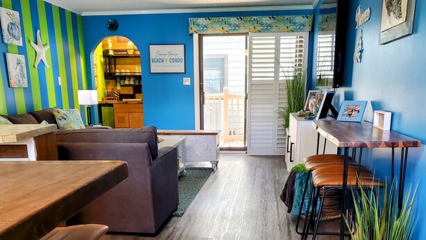A room with a wooden floor, blue walls, a dining area with a table and chairs, a sofa, and a kitchen area with a counter and cabinets. - Exquisite 2 Bedroom/2 Bathroom - Walk to Beach and Boardwalk (Ocean City)