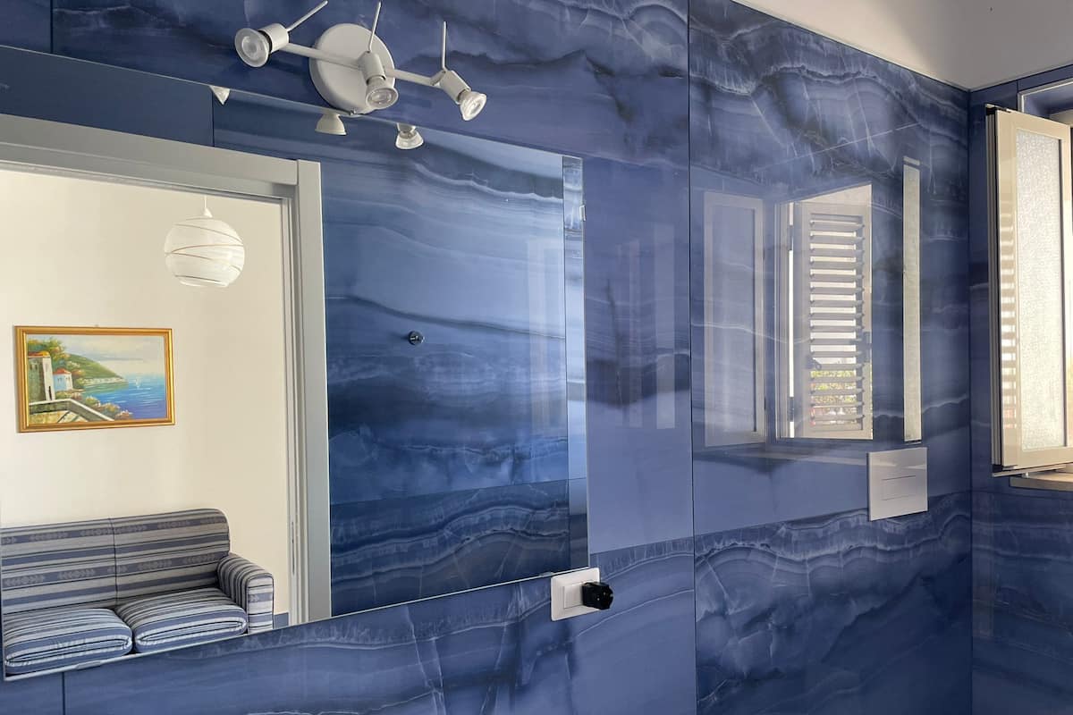 A bathroom with a large mirror, a striped bench, and a painting of a coastal scene.