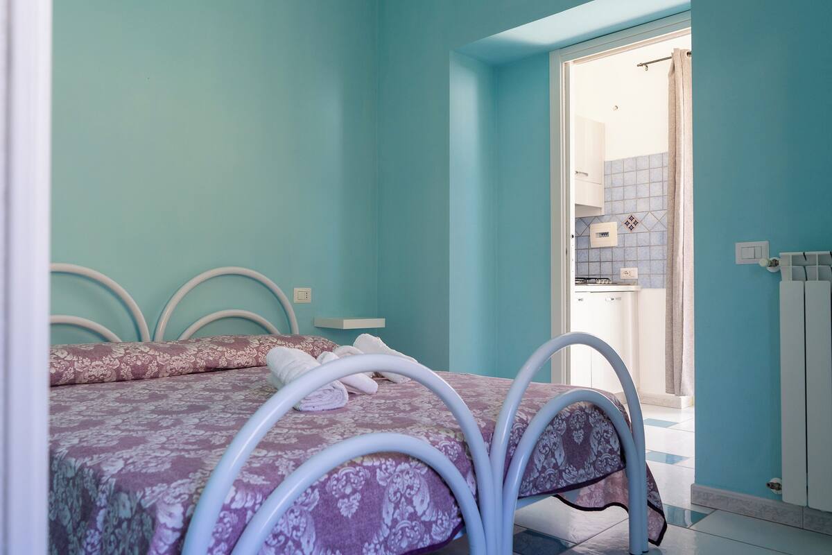 A bedroom with a bed, a bathroom visible through an open door, and a kitchenette area.