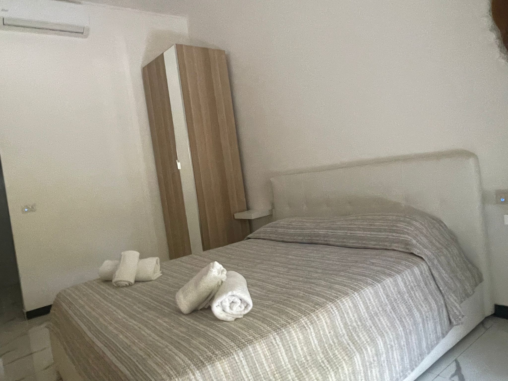 Una cama individual con una colcha a rayas y dos toallas enrolladas. Un armario de madera está colocado contra la pared.