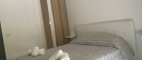 Una cama individual con una colcha a rayas y dos toallas enrolladas. Un armario de madera está colocado contra la pared.