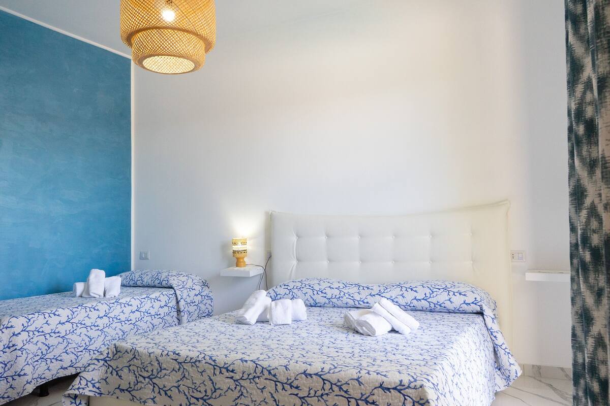 A hotel room with two beds, each with a blue and white patterned bedspread, a white headboard, and a small bedside table with a lamp.