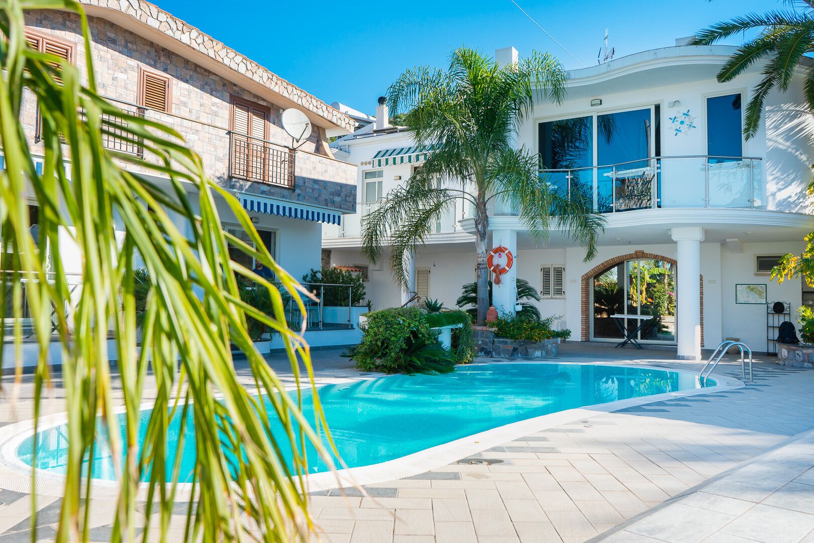 A pool area with a clear blue pool, surrounded by palm trees and a white building with balconies.