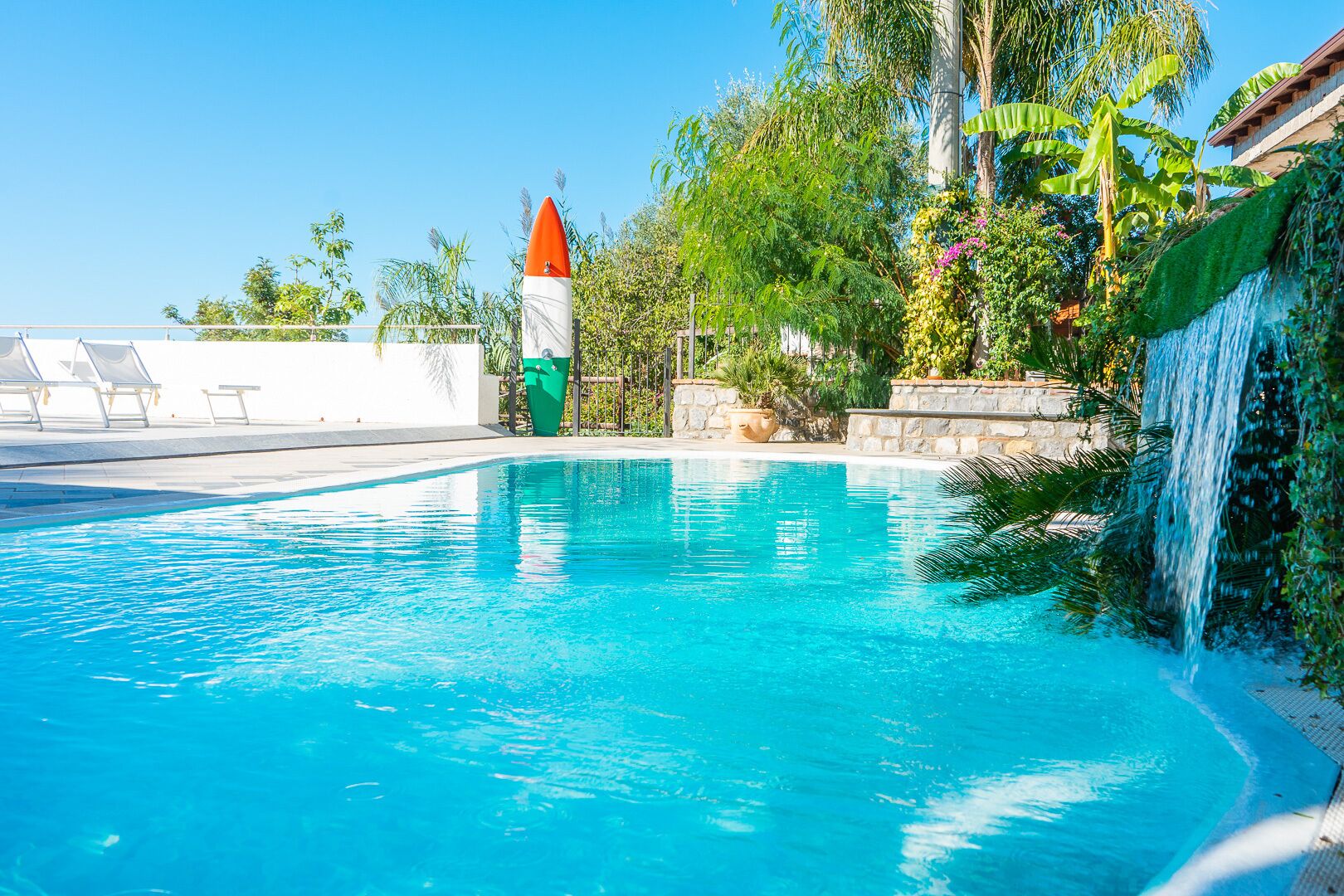 Una piscina con uno scivolo acquatico, circondata da vegetazione rigogliosa e un cielo azzurro.
