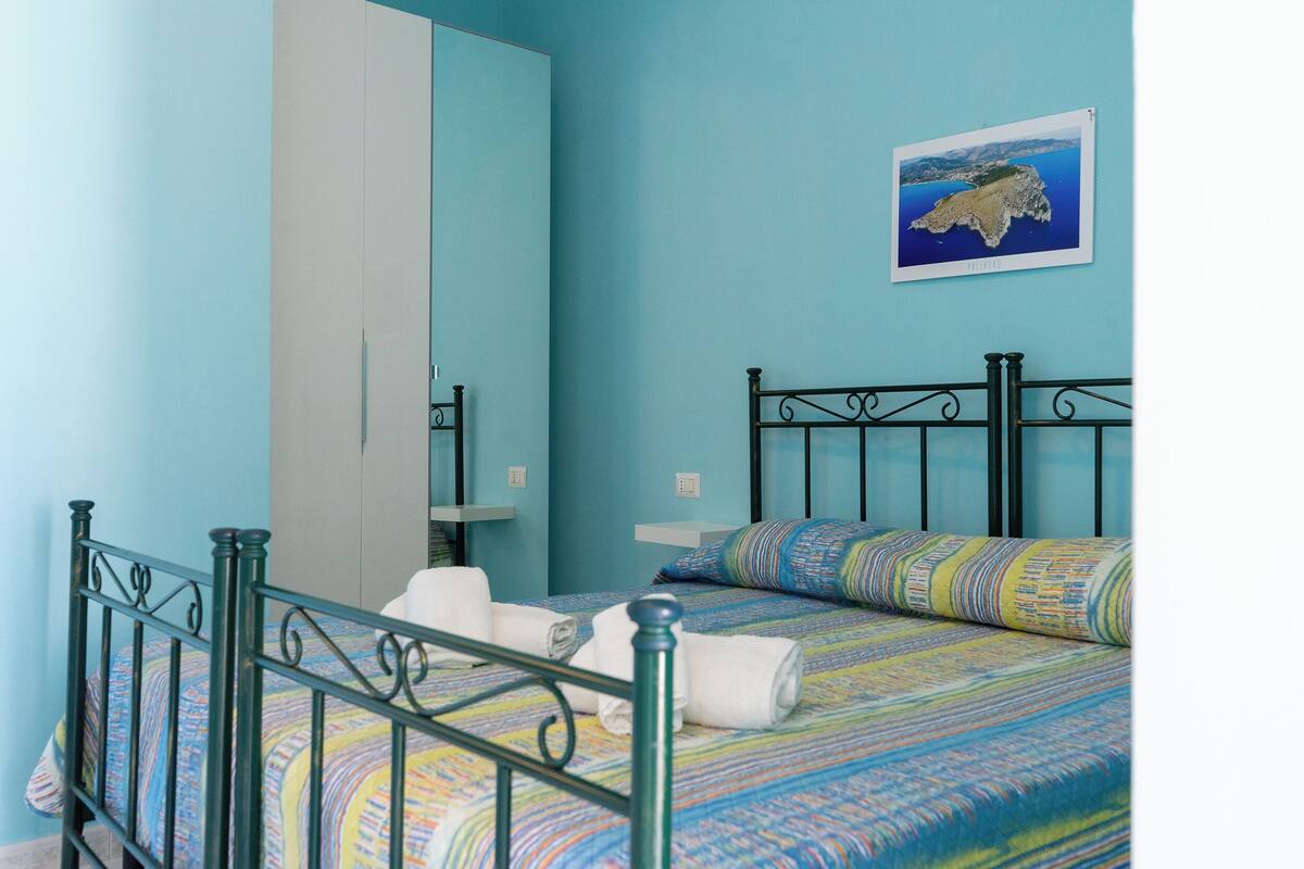 A neatly made bed with a colorful bedspread, a black metal headboard, and a framed picture of a coastal landscape on the wall.