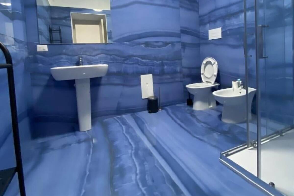 A bathroom with a marble-like blue wall, a pedestal sink, a toilet, and a glass shower enclosure.