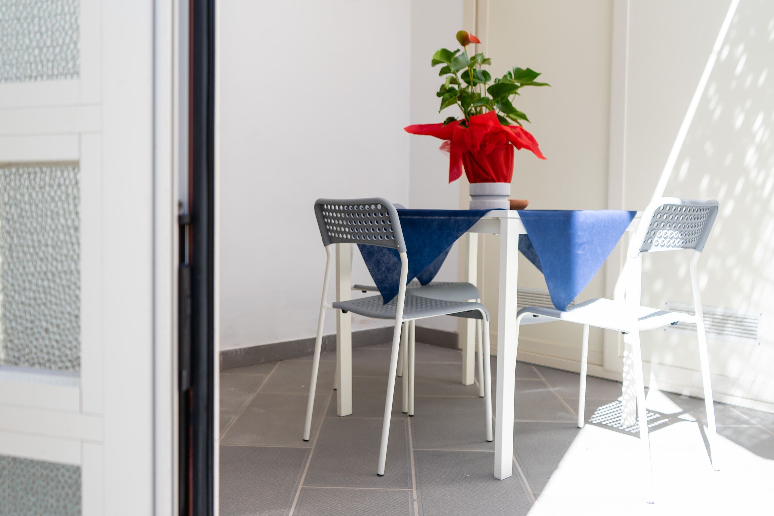 Un balcón con una mesa y dos sillas, una planta en maceta con un lazo rojo y una puerta blanca.