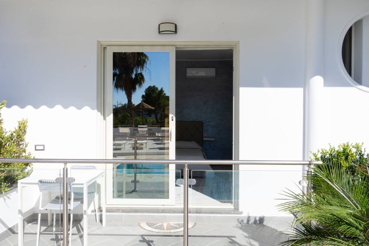 A modern hotel room with a glass door leading to a pool area, a small balcony with a table and chairs, and a view of the pool and palm trees.