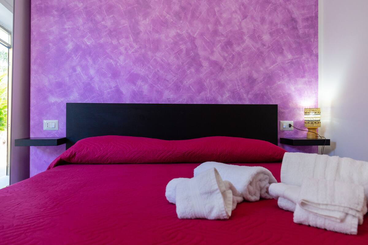 A bed with a red bedspread and pillows, a black headboard, and two white towels on the bed.