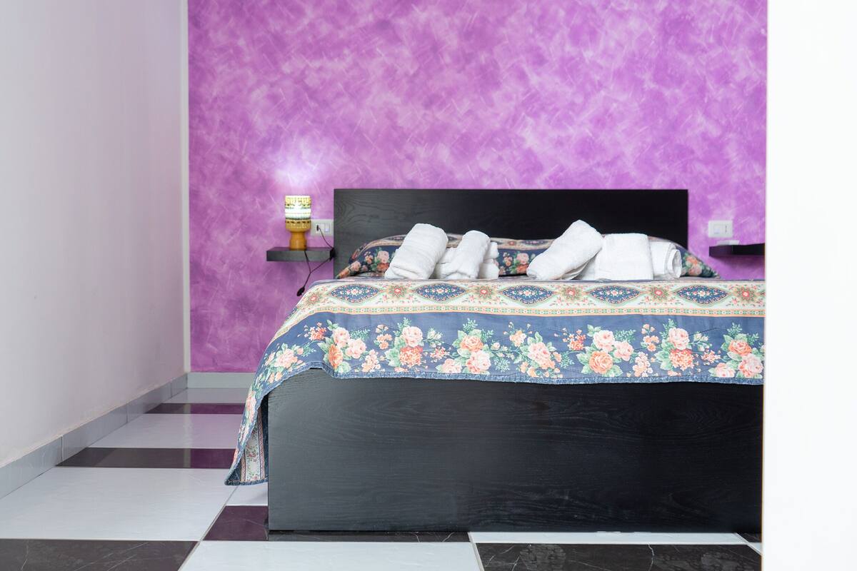 A neatly made bed with floral bedding and towels, a black headboard, and a purple textured wall.