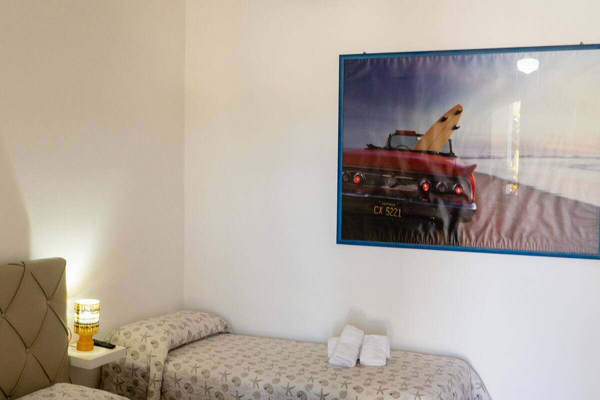 A hotel room with two beds, a ceiling fan, a painting of a car, and a nightstand with a lamp.