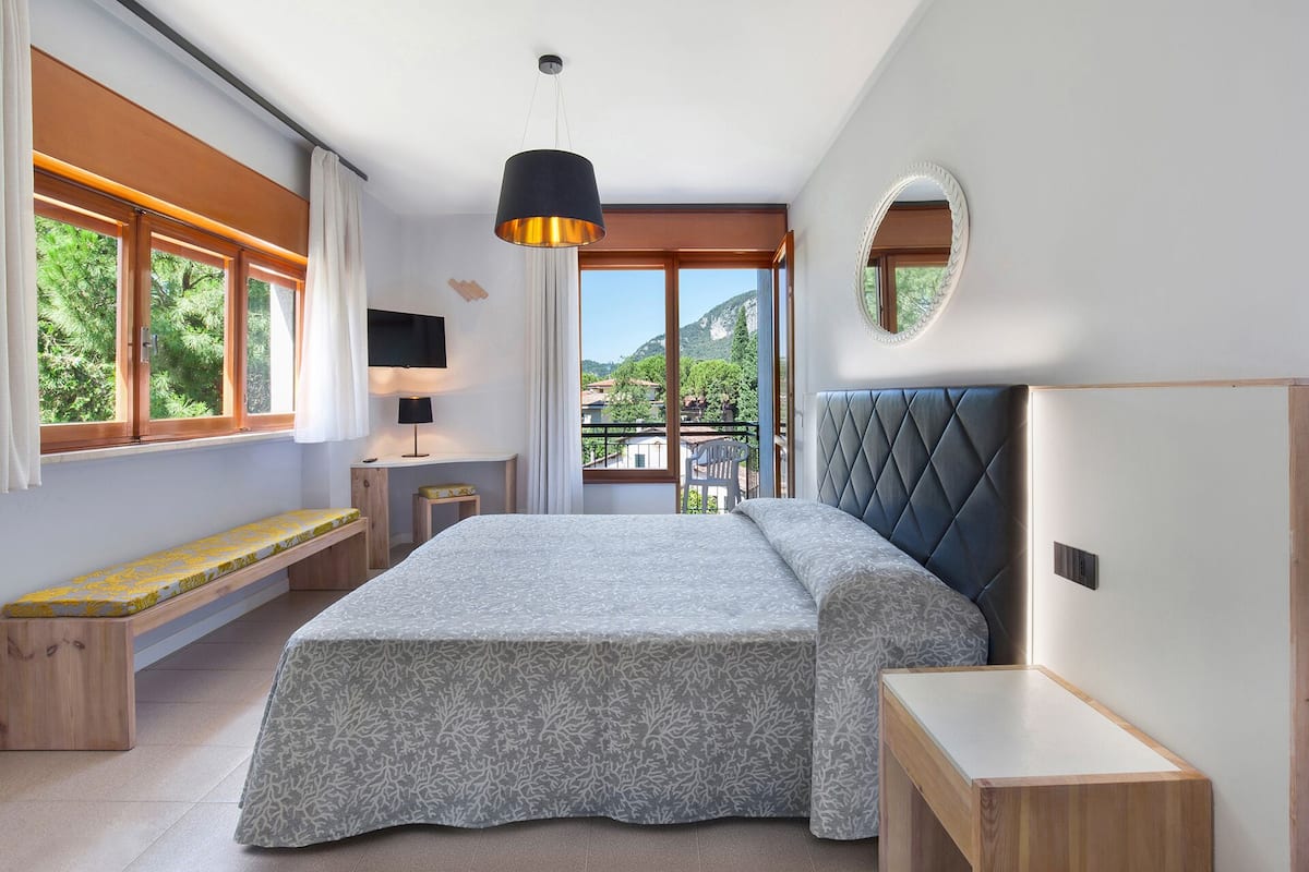 A bedroom with a bed, a desk, a chair, a bench, and a view of the outdoors.