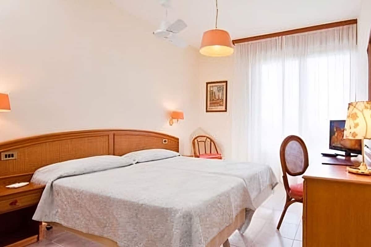 A hotel room with a double bed, a wooden nightstand, a desk with a lamp, a chair, and a television.