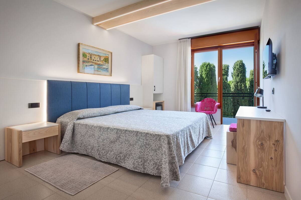 A modern bedroom with a bed, bedside tables, a desk, and a chair. There is a balcony with a view of trees.