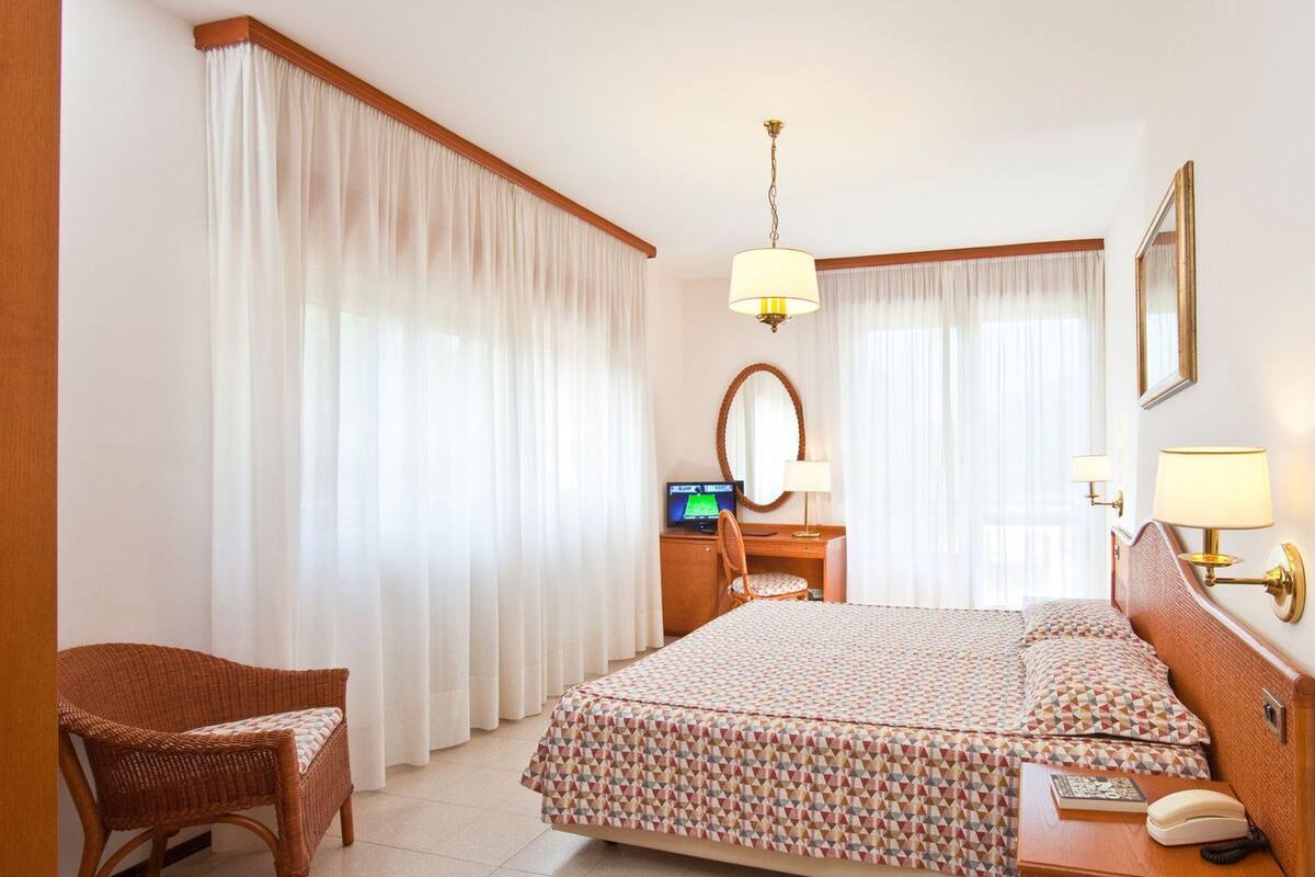 A hotel room with a bed, a desk with a computer, a chair, and a bedside table with a lamp and a phone.