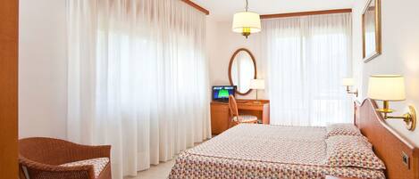 A hotel room with a bed, a desk with a computer, a chair, and a bedside table with a lamp and a phone.