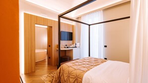 Een moderne hotelkamer met een hemelbed, een flatscreen televisie aan de muur, een klein bureau met stoel, en een aparte badkamer met toilet en wastafel.
