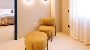 A modern hotel room with a neutral color scheme, featuring a sofa and ottoman set, a wall-mounted light fixture, and a large window with curtains. - Suite Queen Room 2 maggio (Monopoli)