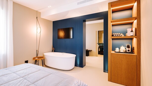 A modern bedroom with a large bed, a freestanding bathtub, and a wooden shelf with toiletries. - Suite Queen Room 2 maggio (Monopoli)