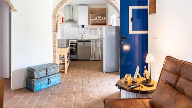 A cozy living room with a brown leather sofa, a blue trunk, and a small wooden table with a lamp. A kitchen with a blue door and an archway is visible in the background. - Sunrise Monsaraz Blue - Casa com 2 Quartos Jardim (Évora)