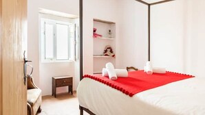 A bedroom with a four-poster bed, a small bedside table, a nightstand, a window, and a shelf with a doll. - Sunrise Monsaraz Blue - Casa com 2 Quartos Jardim (Évora)