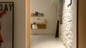 Un pasillo estrecho con suelo blanco, marco de puerta de madera, una silla blanca, un escritorio con lámpara y una pared de piedra.