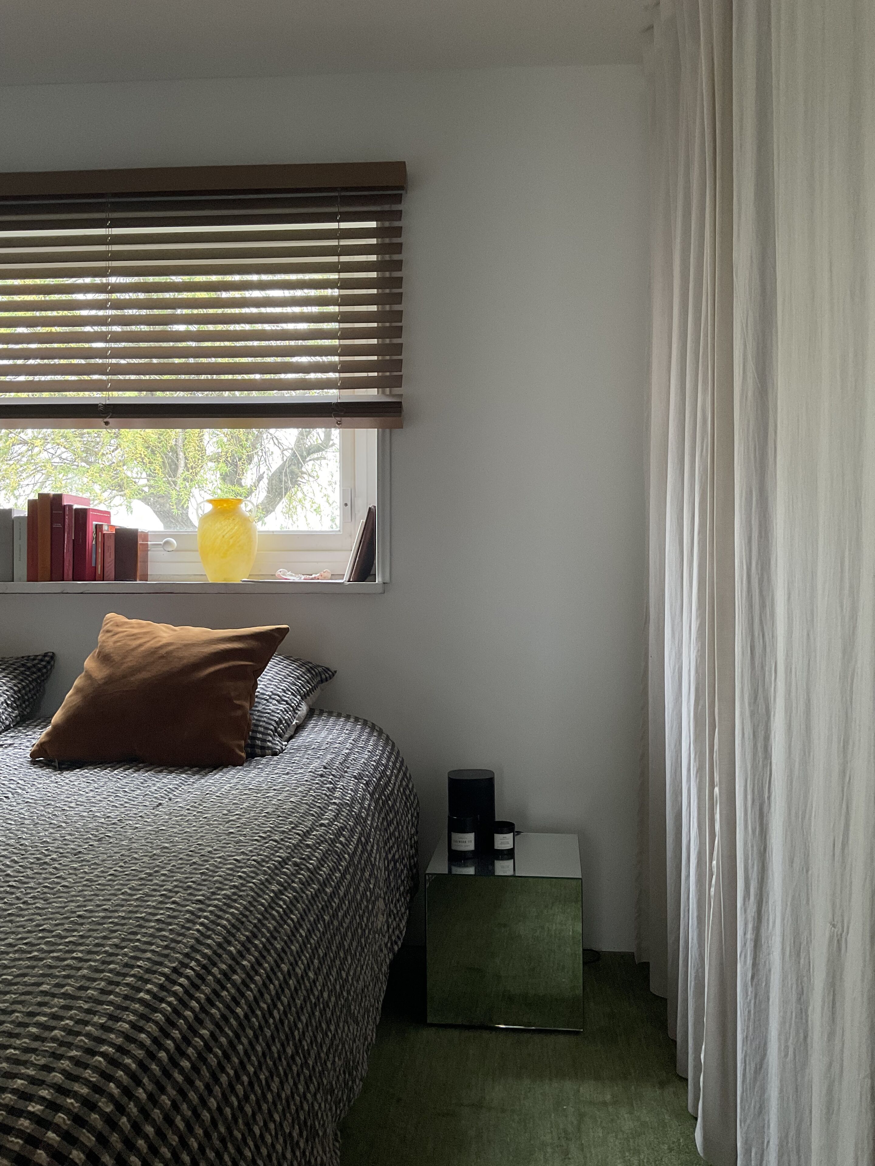 Une chambre à coucher avec un lit, une fenêtre avec des stores, une table de chevet avec une lampe et un rideau.