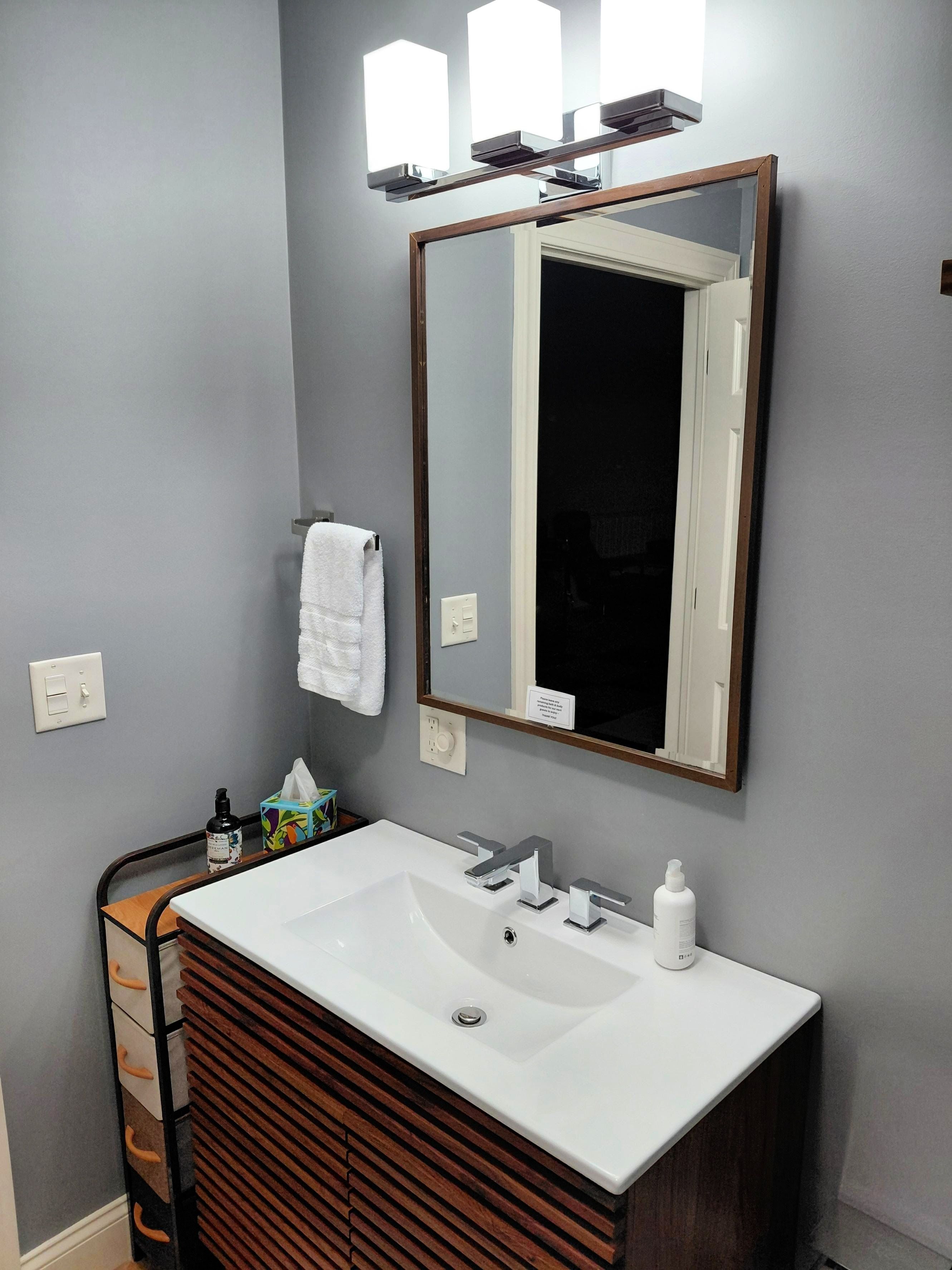 A bathroom with a white sink, wooden vanity, a mirror with lights, and a towel hanging on the wall.