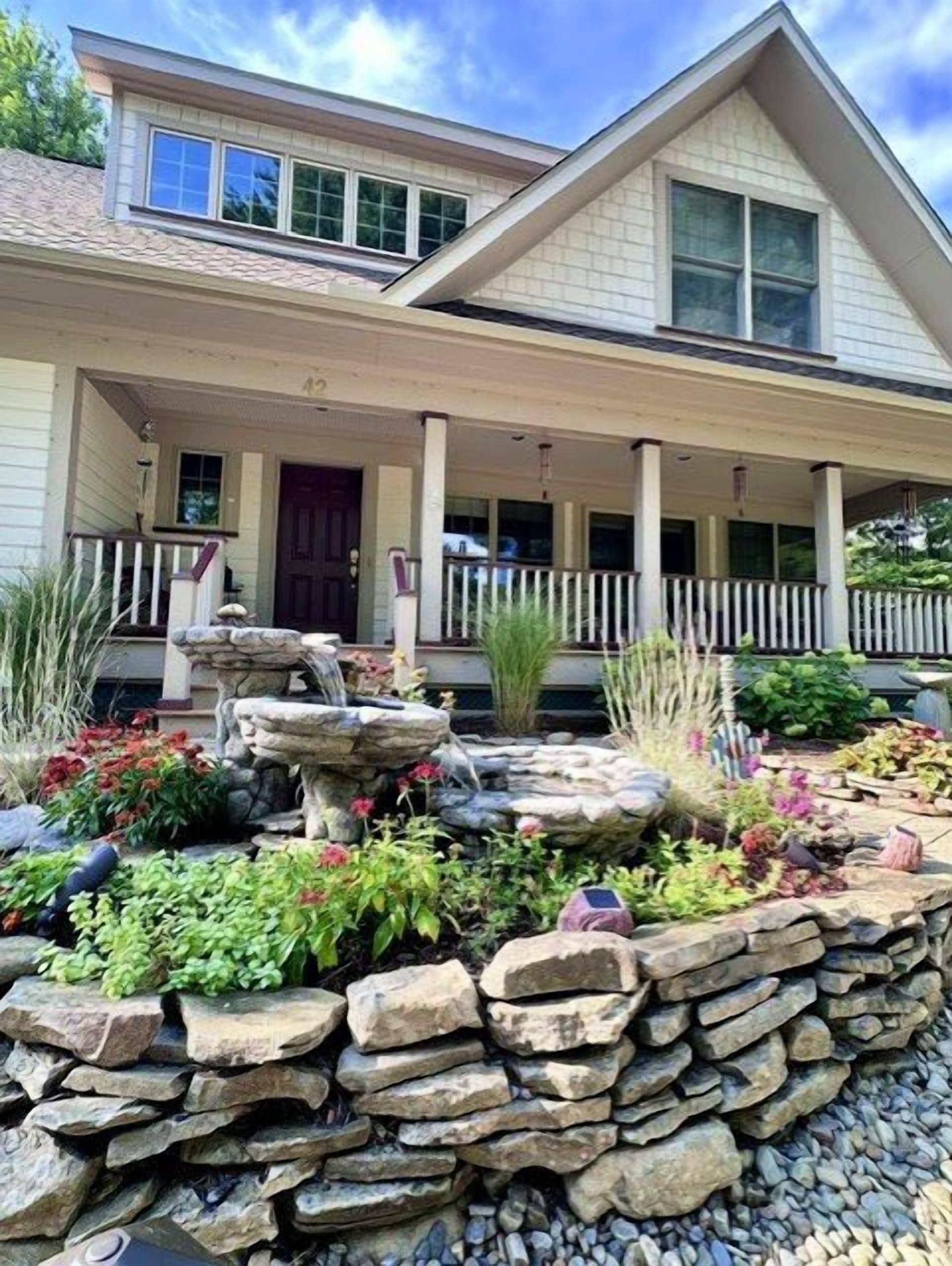 A two-story house with a front porch, a stone retaining wall, and a small water feature.