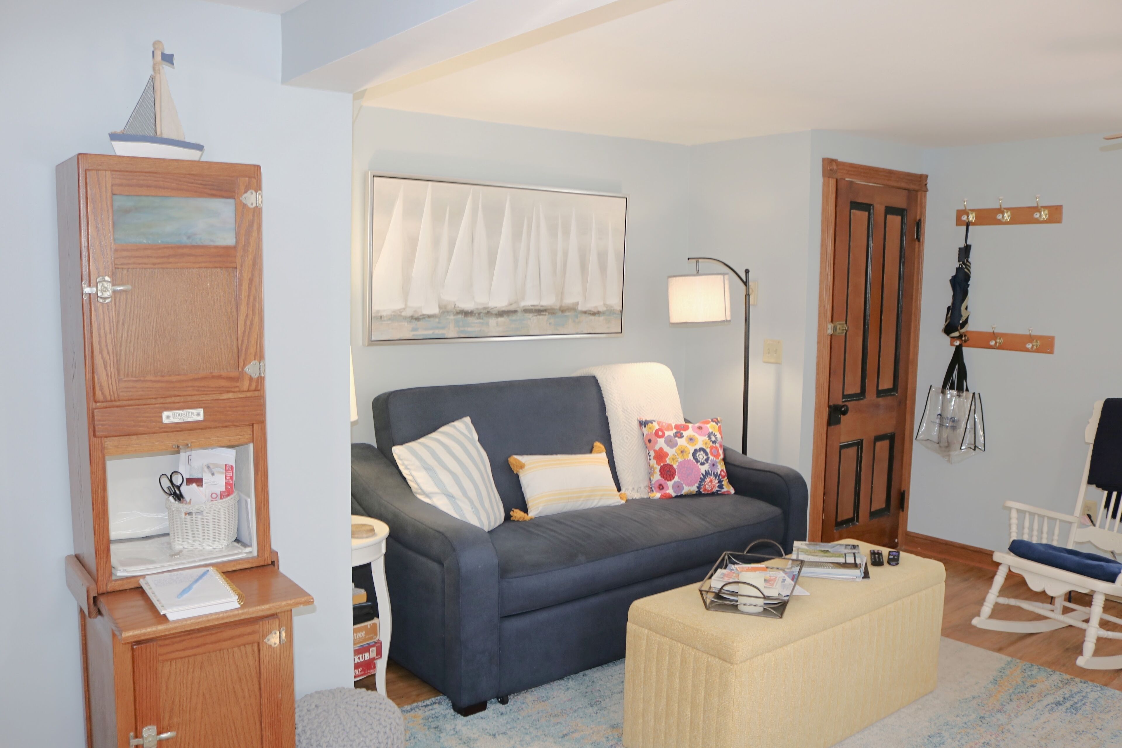 A living room with a sofa, a small wooden cabinet, a rocking chair, a lamp, and a painting of sailboats.