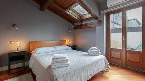 Une chambre avec un grand lit, des poutres apparentes en bois au plafond, un puits de lumière et une vue sur l’extérieur grâce à une porte vitrée.