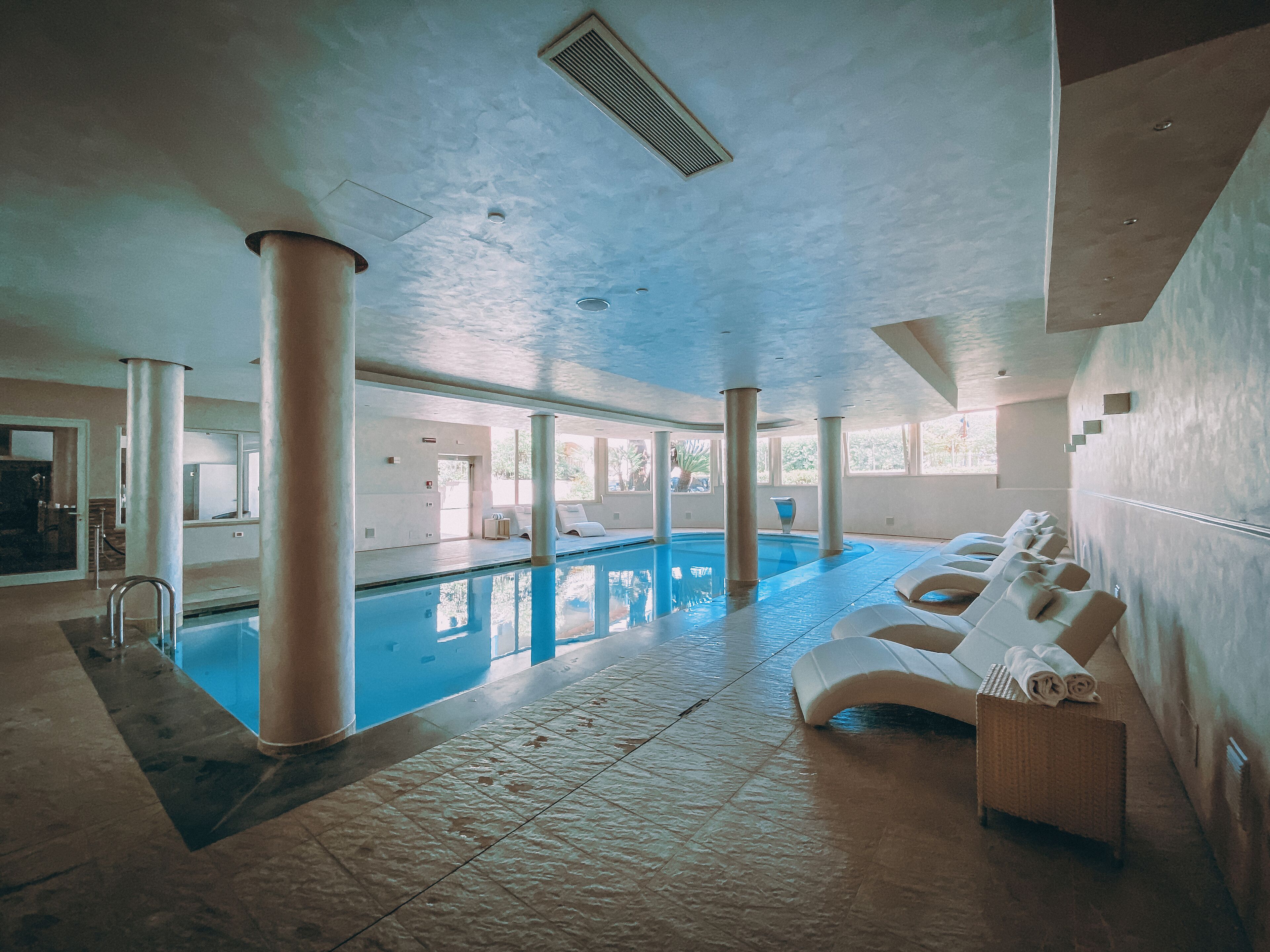 An indoor swimming pool with lounge chairs and a sunbed, surrounded by large windows and columns.