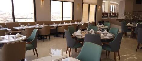 Un restaurante con mesas dispuestas para comer, ventanas amplias y vistas a la ciudad.
