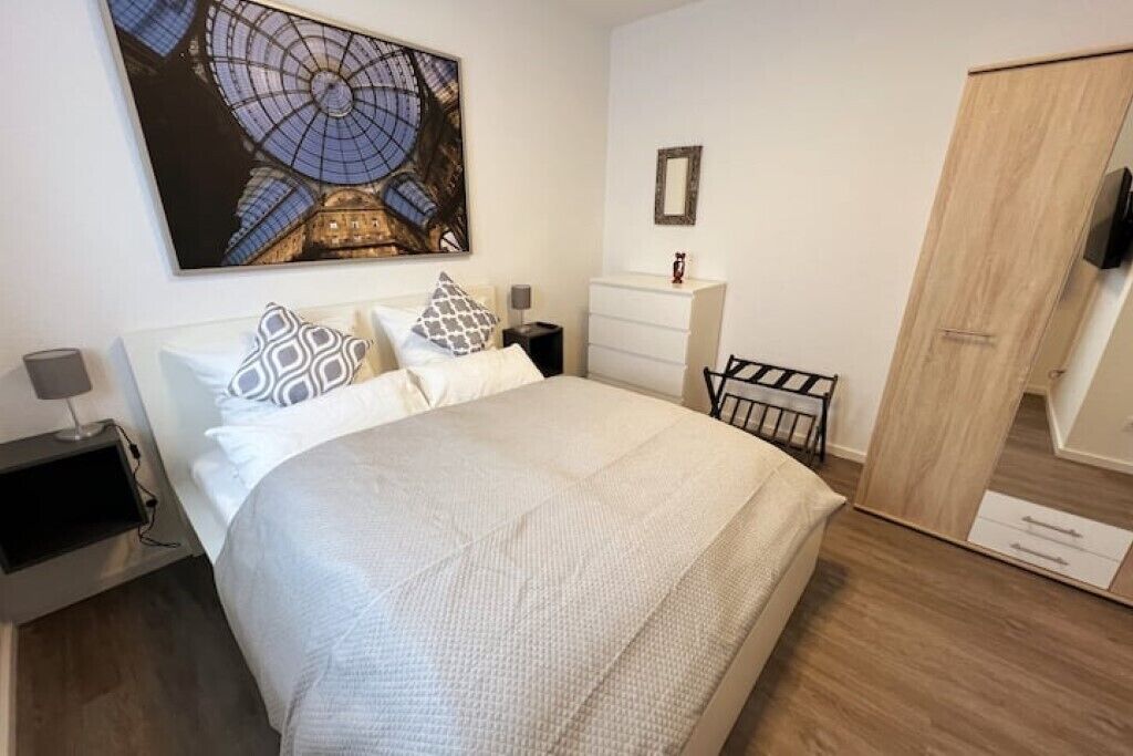 Une chambre à coucher avec un grand lit, une table de chevet, une commode, un miroir et une œuvre d’art accrochée au mur.