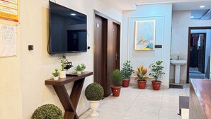 A modern hotel lobby with a flat-screen TV, wooden reception desk, potted plants, and a ceiling fan. - Hotel Raghuveera Palace (Ujjain)