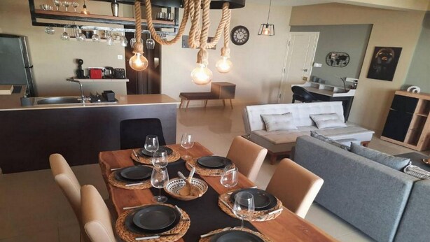 A modern kitchen with a central island, dining area with a wooden table set for four, and a grey sofa in the living area. - Paramount Gardens Resorts C201 (Tersefanou)