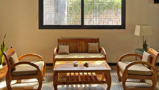 A room with a wooden ceiling, a large window with a view of greenery, a sofa, two wooden chairs, a coffee table, and a hanging light fixture.