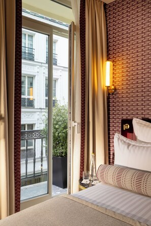 A hotel room with a large sliding glass door leading to a balcony, a bed with white linens, a wall-mounted light fixture, and a small table with a glass and a bottle.