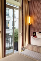 Et hotelværelse med en stor skydedør, der fører ud til en balkon, en seng med hvide sengetøj, en vægmonteret lampe og et lille bord med et glas og en flaske.