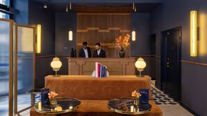 A reception area with a wooden reception desk, two standing receptionists, a check-in counter, and a check-in desk with a sofa. There are two round tables with lamps and a vase of flowers. - Hôtel Filigrane & Spa (Paris)