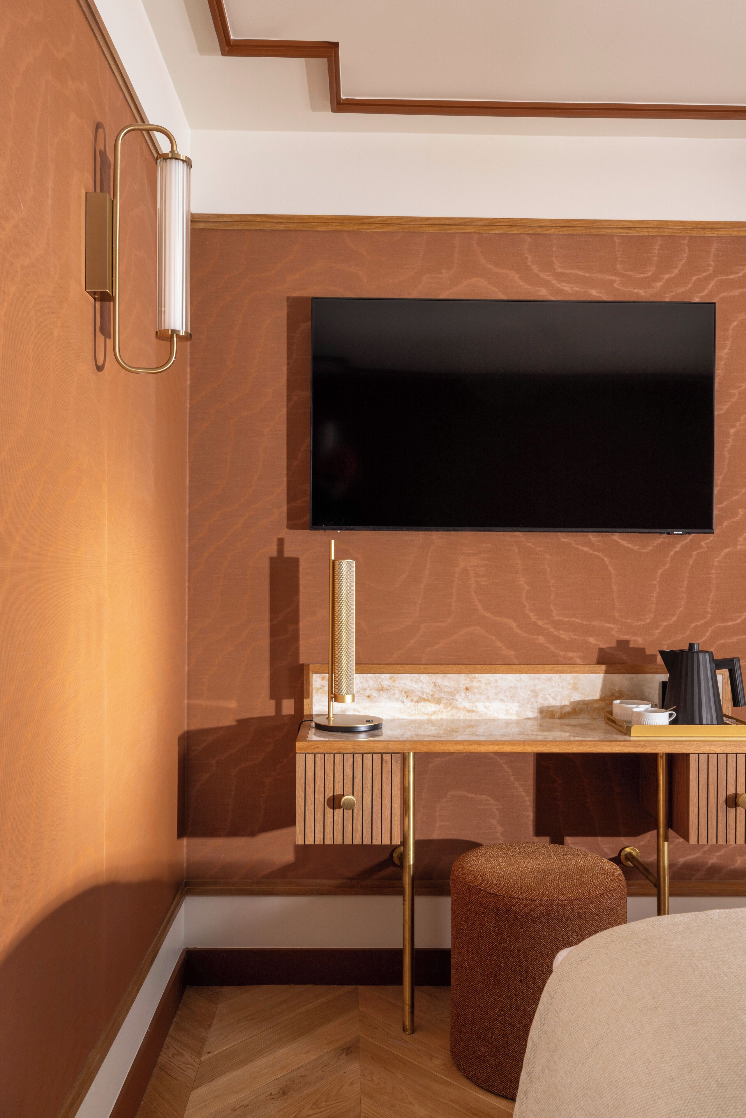A modern hotel room with a flat-screen TV mounted on the wall, a desk with a lamp, and a brown ottoman.