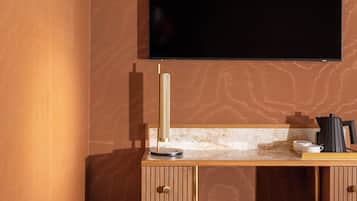 Habitación de hotel moderna con una televisión de pantalla plana montada en la pared, un escritorio con una lámpara y un otoman marrón.