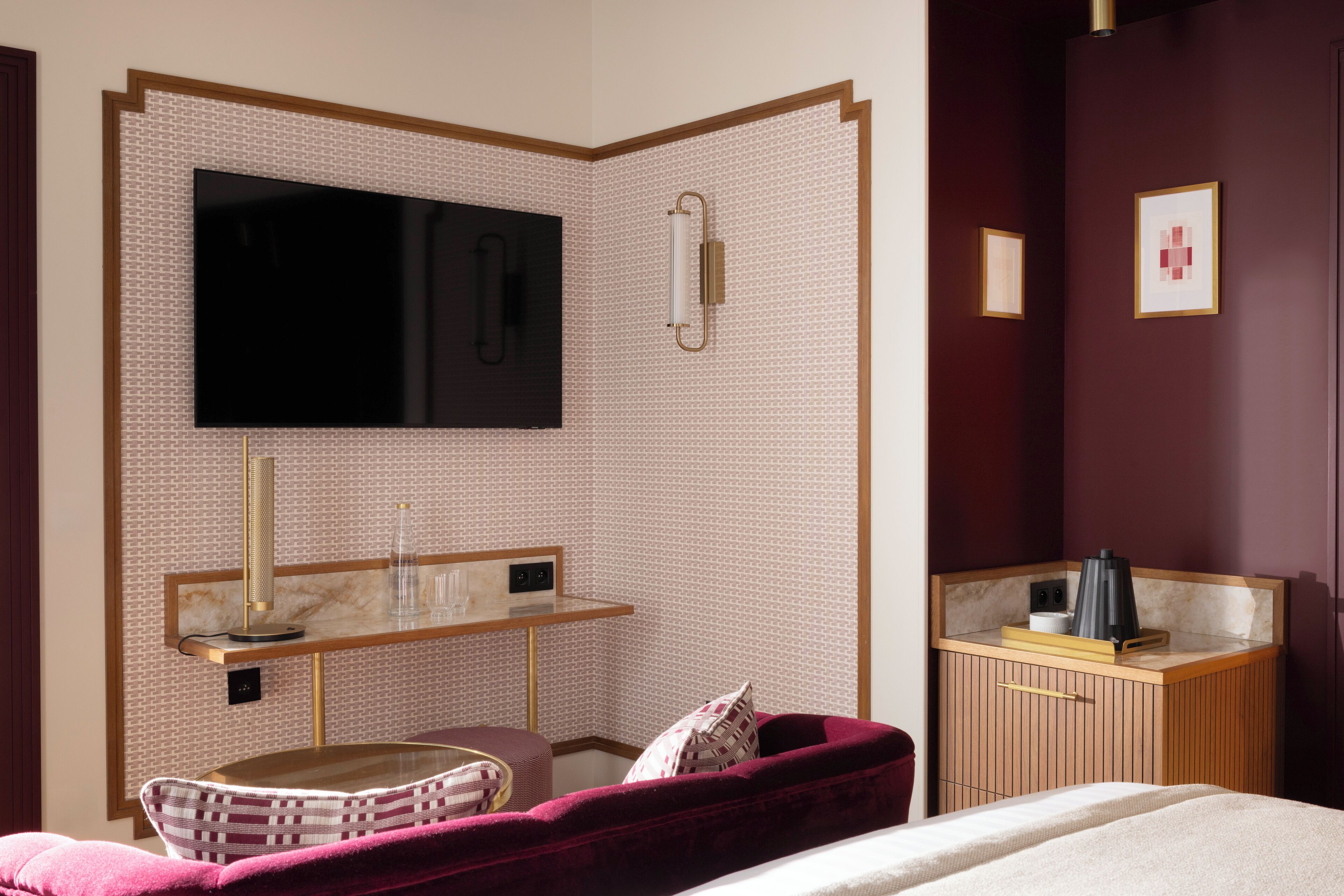 A hotel room with a large flat-screen TV mounted on a patterned wall, a bedside table with a lamp, a cushioned bench, and a wooden cabinet with a mirror.