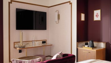 A hotel room with a large flat-screen TV mounted on a patterned wall, a bedside table with a lamp, a cushioned bench, and a wooden cabinet with a mirror.