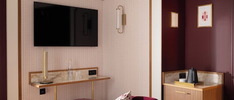 Une chambre d’hôtel dotée d’un grand téléviseur à écran plat fixé sur un mur orné de motifs, d’une table de chevet avec une lampe, d’un banc rembourré et d’une armoire en bois avec un miroir.