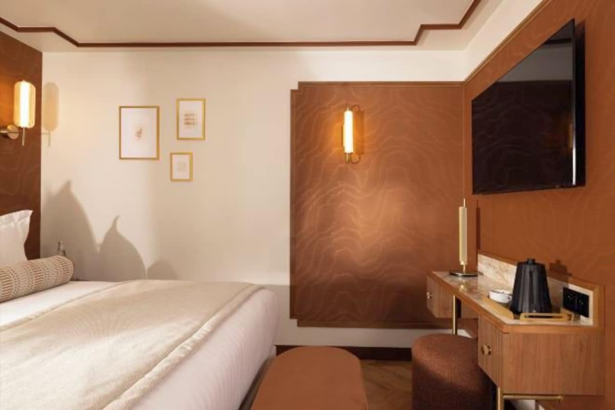 A hotel room with a bed, a desk with a lamp, a flat-screen TV, and framed pictures on the wall.