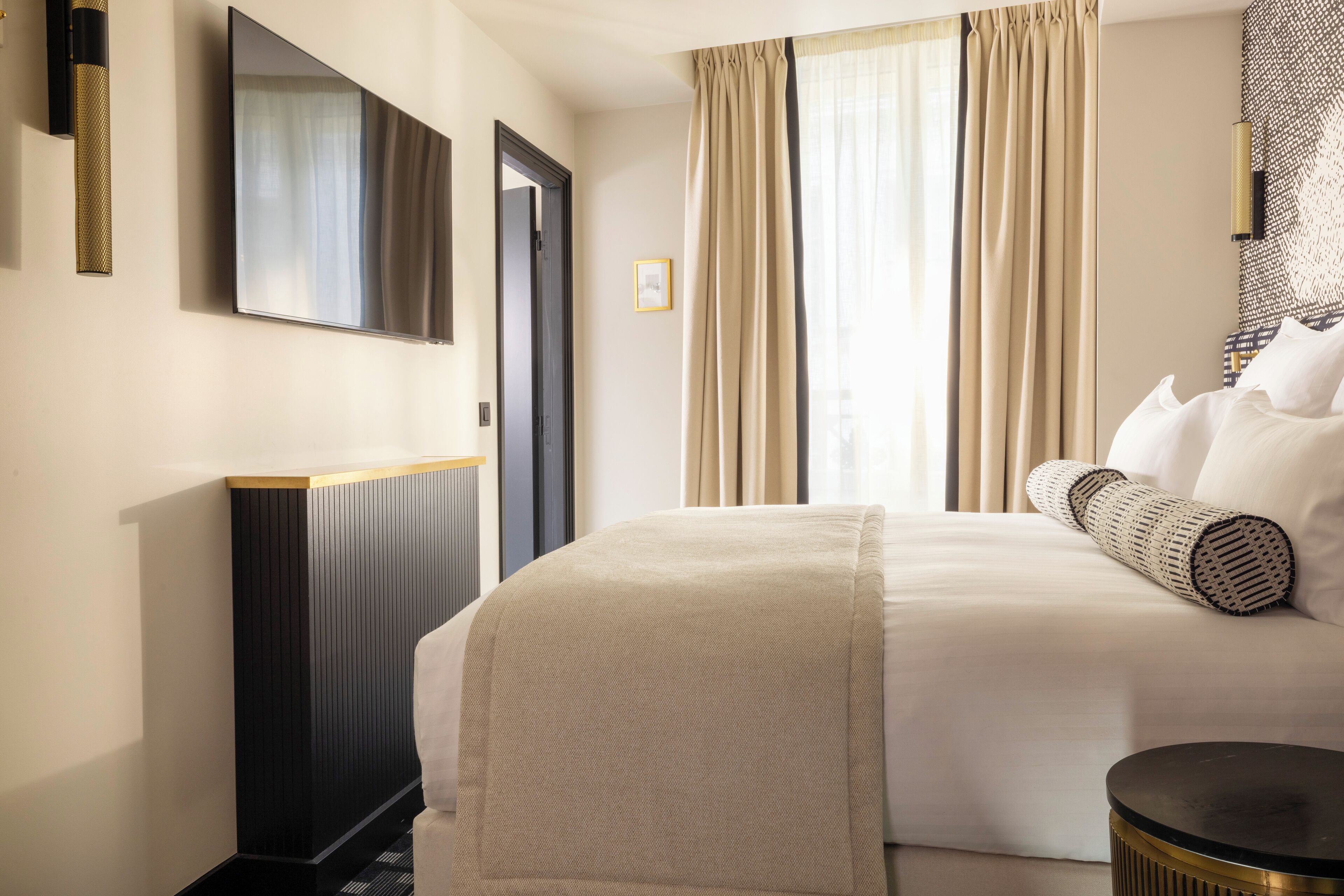 Une chambre d’hôtel avec un lit, une petite table d’appoint ronde et noire, un téléviseur fixé au mur et une grande fenêtre avec des rideaux.