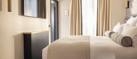 Habitación de hotel con cama, una mesita redonda negra, televisor montado en la pared y una ventana grande con cortinas.