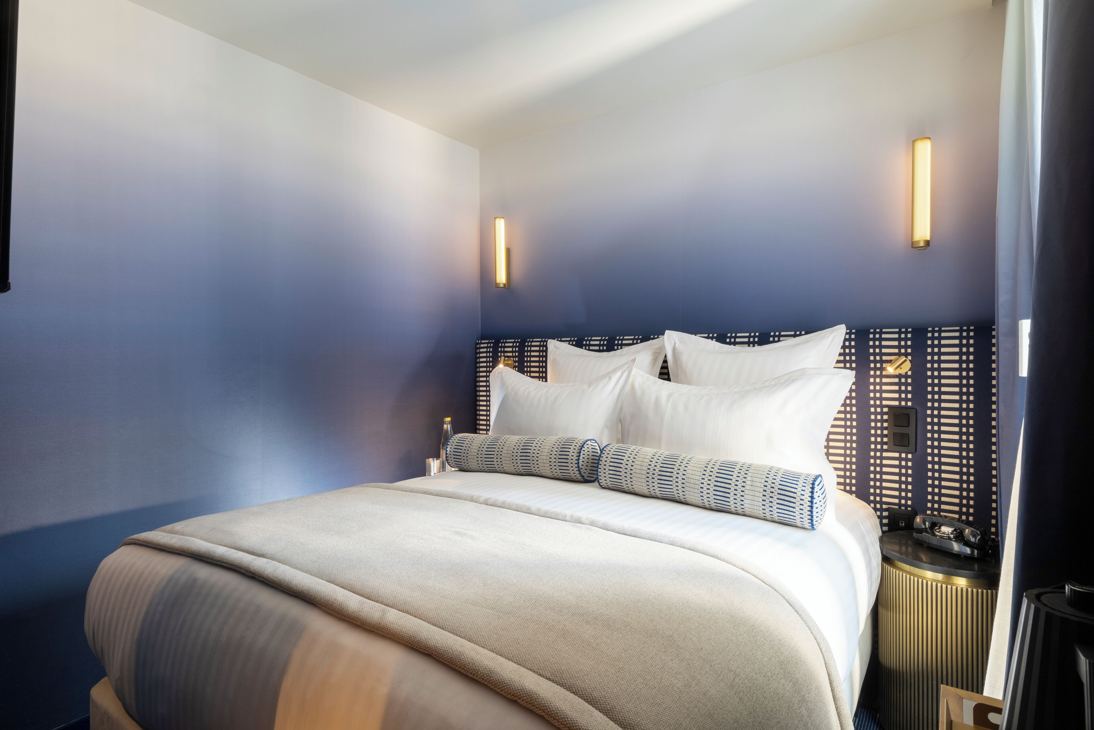 Una cama bien hecha con cabecera, una mesita de noche con lámpara y un aplique de pared.