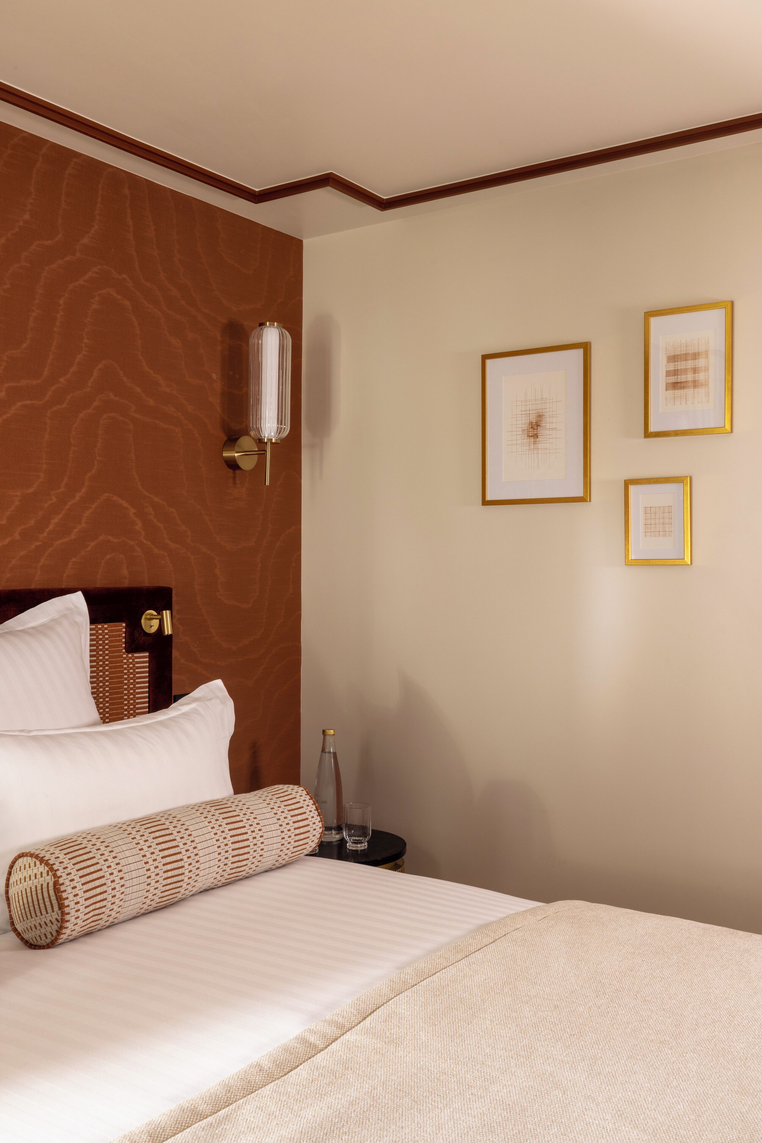 A neatly made bed with a rolled-up blanket, a bedside table with a bottle and glasses, and framed artwork on the wall.